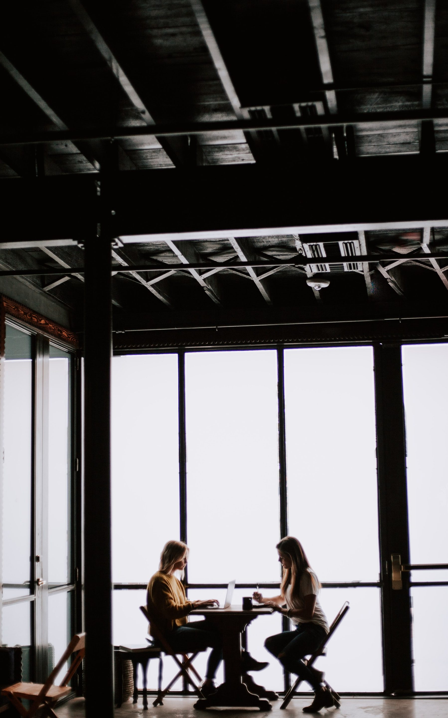 photo of two women facing each other sitting in front of table near glass wall
